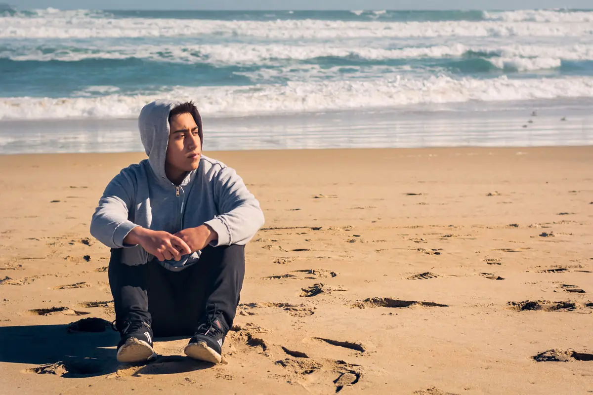 Nachdenklicher junger Mann sitzt auf dem Sand am Strand, nachdem seine Ex-Freundin einen neuen Freund hat.