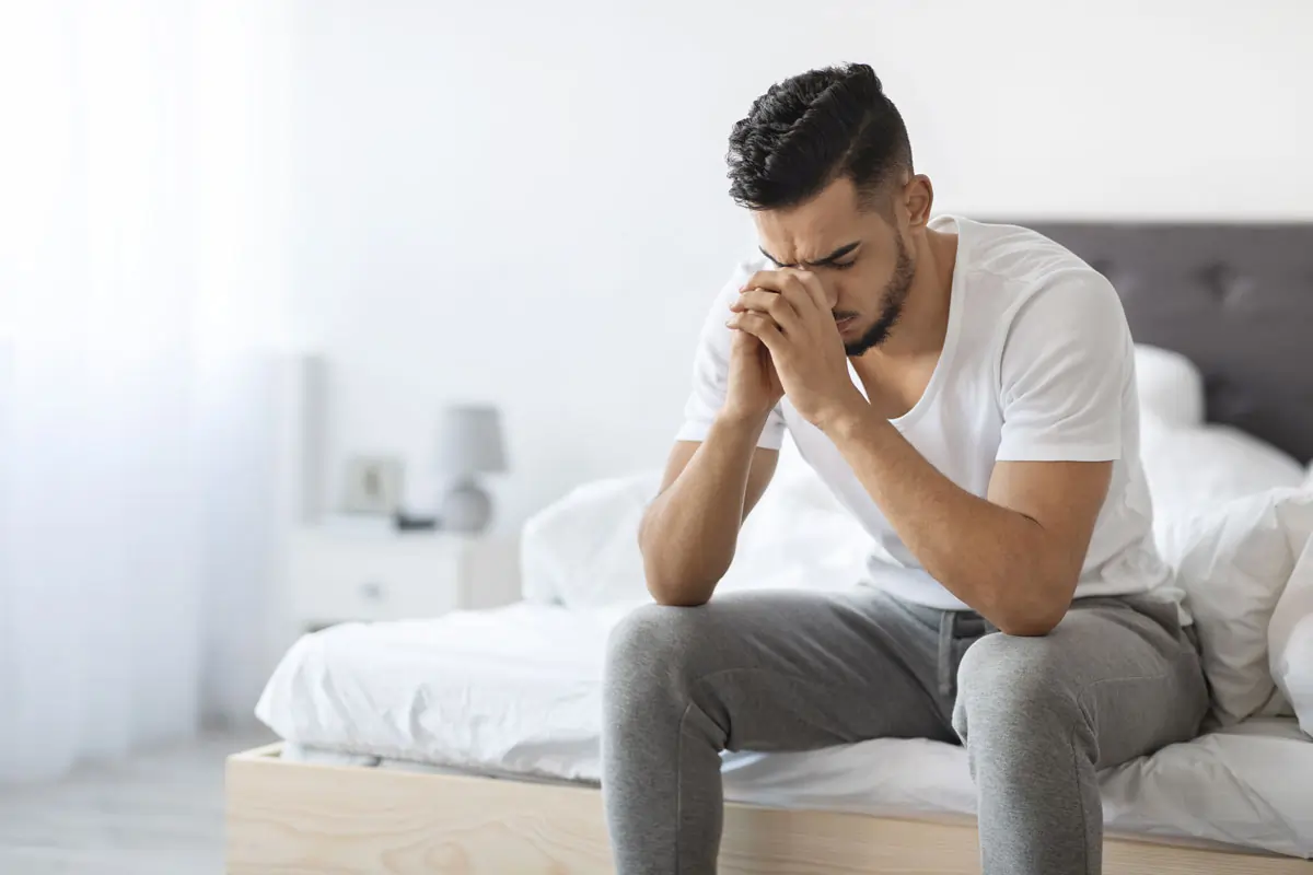Ein nachdenklicher und trauriger Mann sitzt auf einem Bett, symbolisiert den emotionalen Schmerz nach einem Vertrauensbruch in der Beziehung, wenn der Partner betrügt.