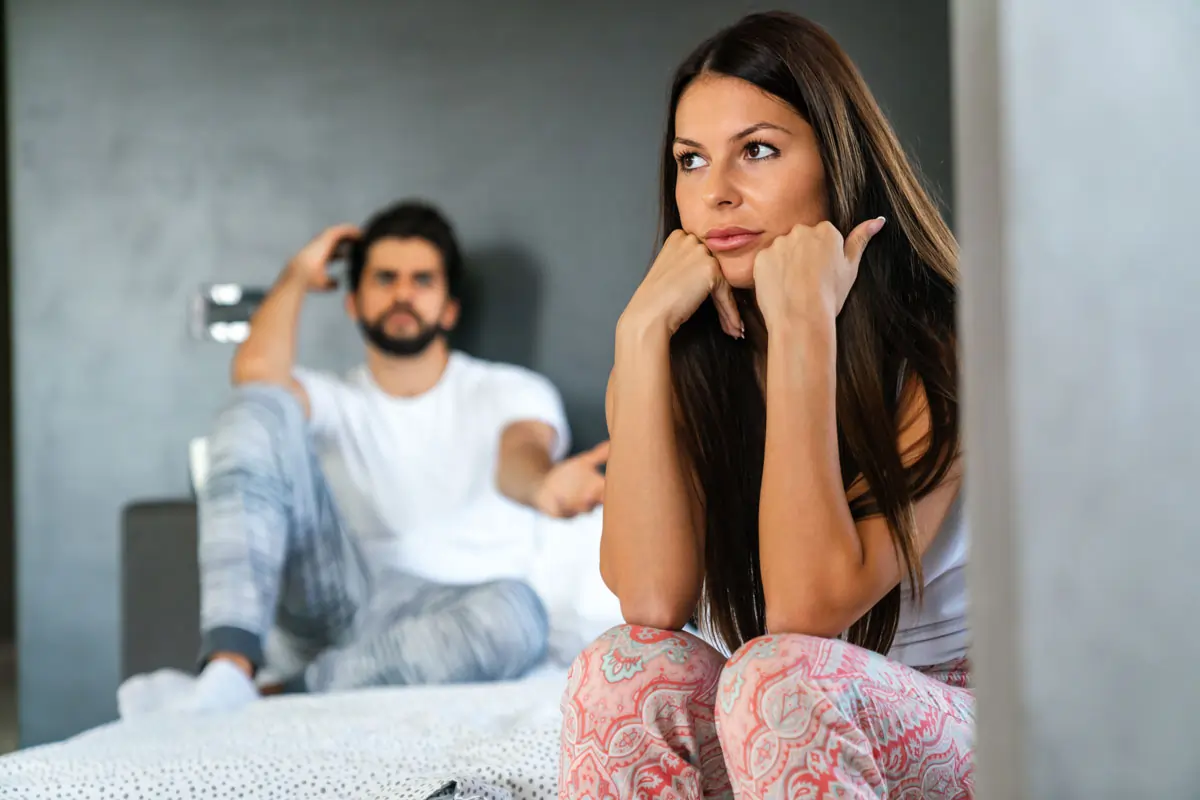 Eine nachdenkliche Frau sitzt im Vordergrund, während ihr Partner distanziert im Hintergrund ist – symbolisiert emotionale Distanz, wenn ein Mann sich komisch verhält