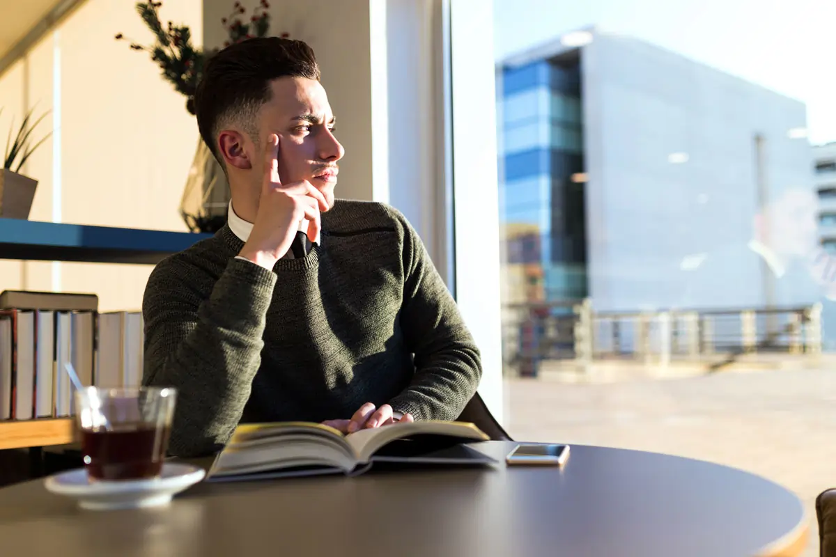 Ein nachdenklicher Mann sitzt allein an einem Tisch und schaut aus dem Fenster, was emotionale Symptome von Liebeskummer darstellt.