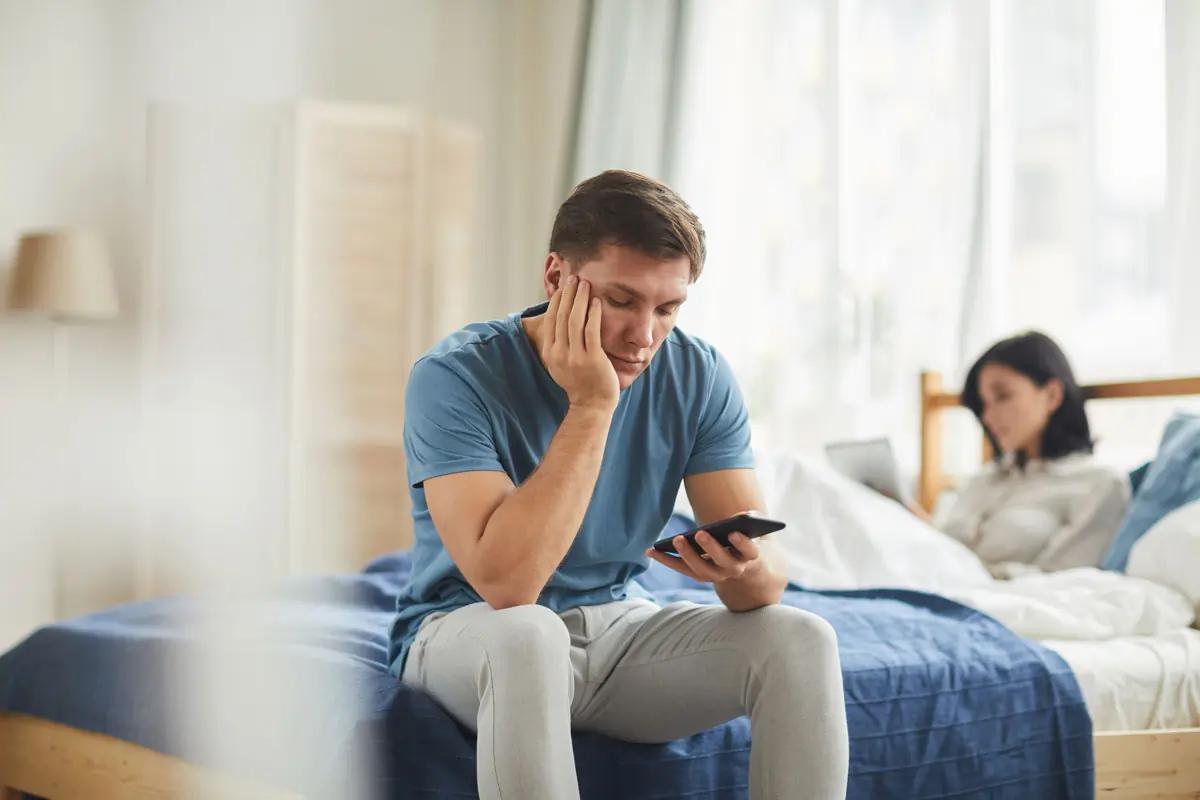Ein nachdenklicher Mann sitzt mit einem Smartphone in der Hand auf dem Bett, während eine Frau im Hintergrund beschäftigt ist. Das Bild symbolisiert emotionale Distanz und Beziehungsprobleme, die durch Depressionen und Untreue entstehen können.